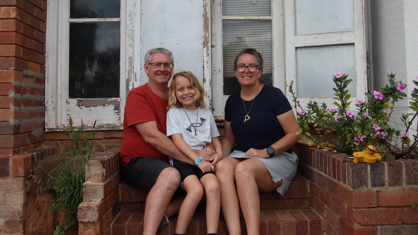 A man, woman, and child, sit on the stairs of a brick house with wooden doors.