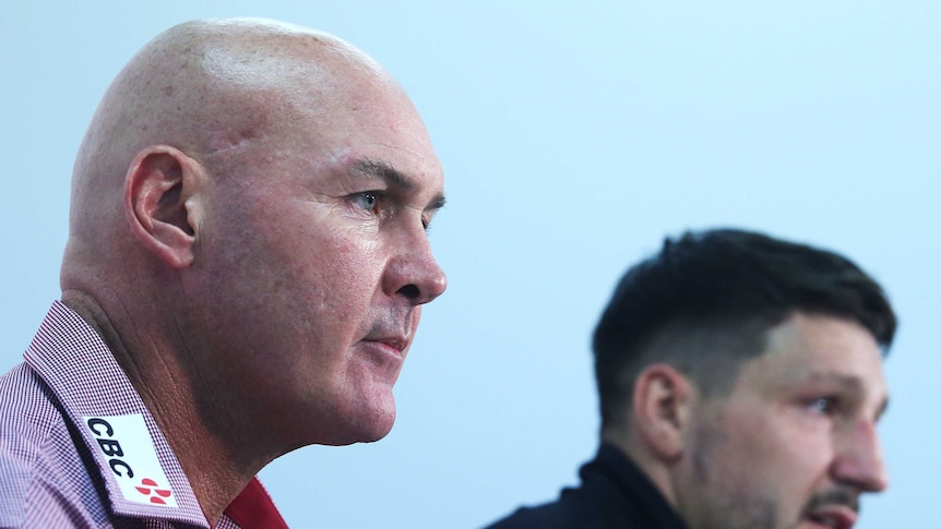 An older bald man and a younger, more hirsute fellow sit together at what appears to be a press conference.