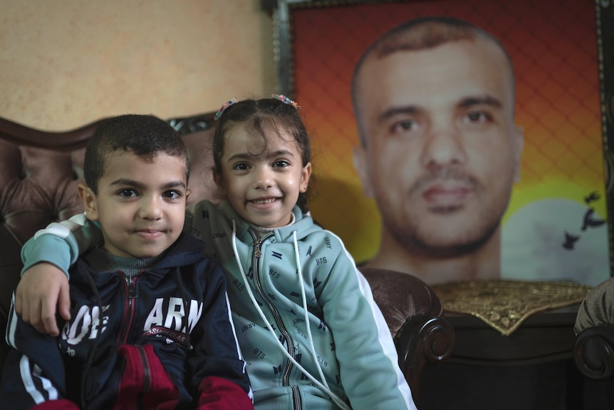 A little boy and girl sit next to each other on an ornate armchair in front of a portrait of a young man. They are both smiling