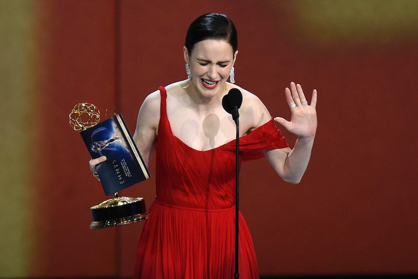 A woman in a red evening gown stands in front of a microphone holding an award