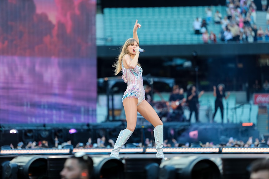 Taylor Swift throws a hand in the air as she sings on stage in a sparkling bodysuit.