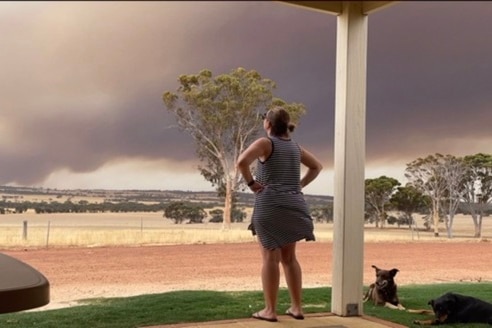 A woman on a rural property stands on her porch looking out at dark smoke on the horizon.