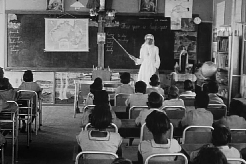 Historical photo shows a white teacher at the front of a class of aboriginal children pointing at a map of Australia.