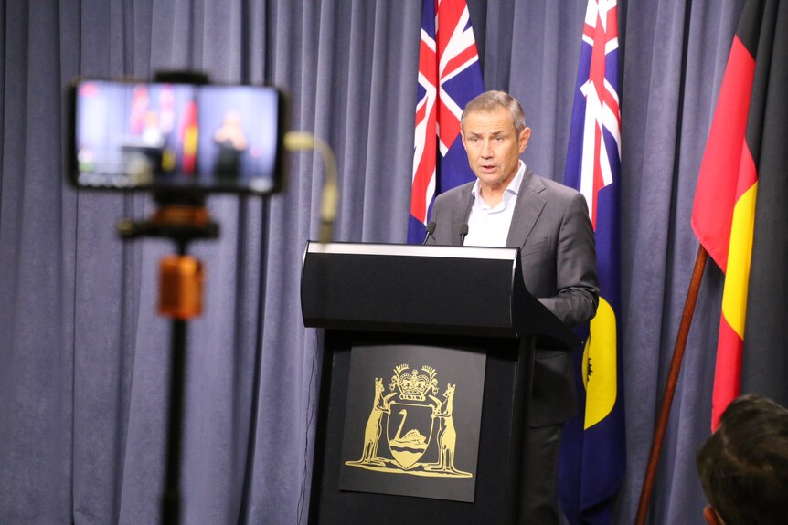 Roger Cook stands at a podium against a blue curtain and flags, with an iphone out of focus in the foreground.
