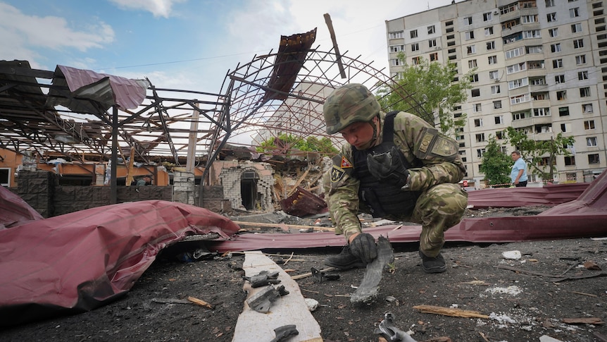 A soldier squats down to inspect fragments of a bomb surrounded by debris following an attack