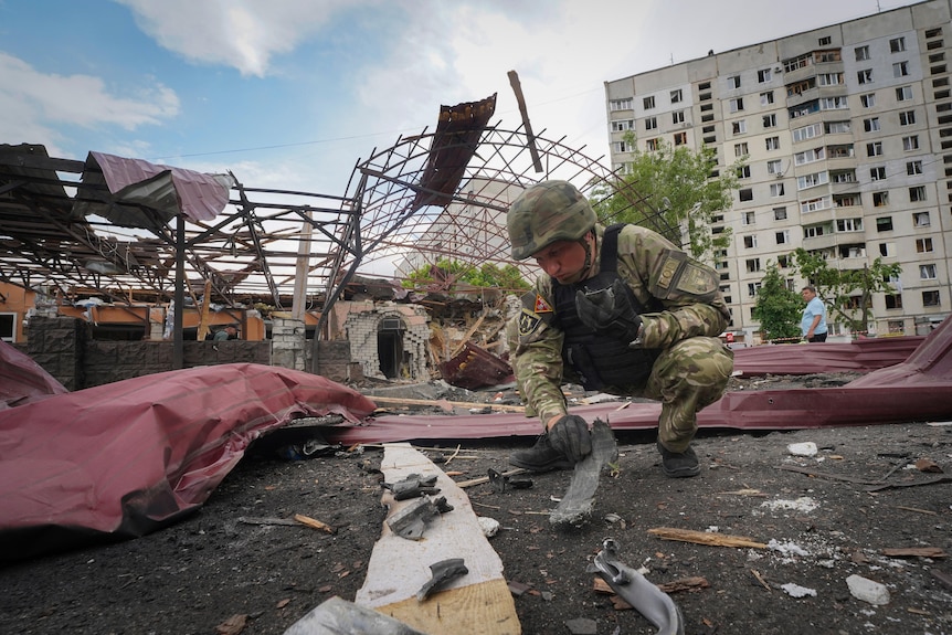 A soldier squats down to inspect fragments of a bomb surrounded by debris following an attack