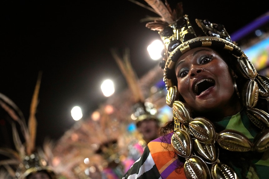 a woman in a shiny headress opens her mouth and eyes up wide as she dances and looks toward the camera at Carnival