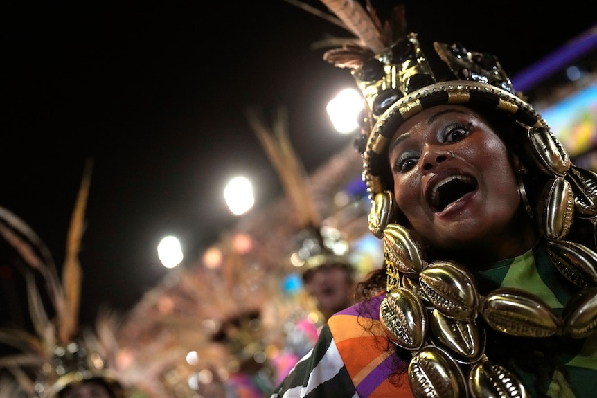 une femme dans une coiffe brillante ouvre la bouche et les yeux grands ouverts alors qu'elle danse et regarde vers la caméra au carnaval