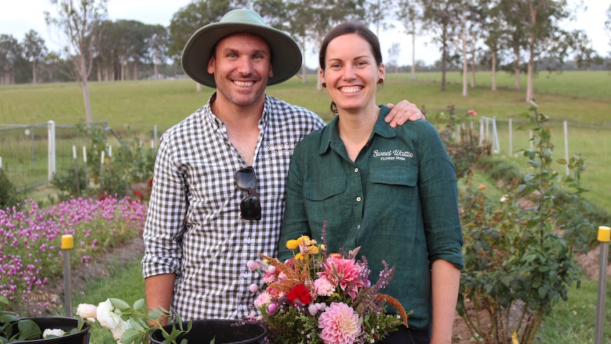 A man in a blue and white check shirt stands next to a lady wearing a green shirt on a flower farm.