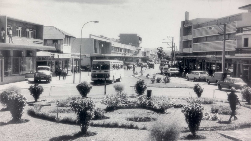 Wide shot of a street scene. A roundabout in the foreground, a bus approaching it and cars, buildings and people on either side.