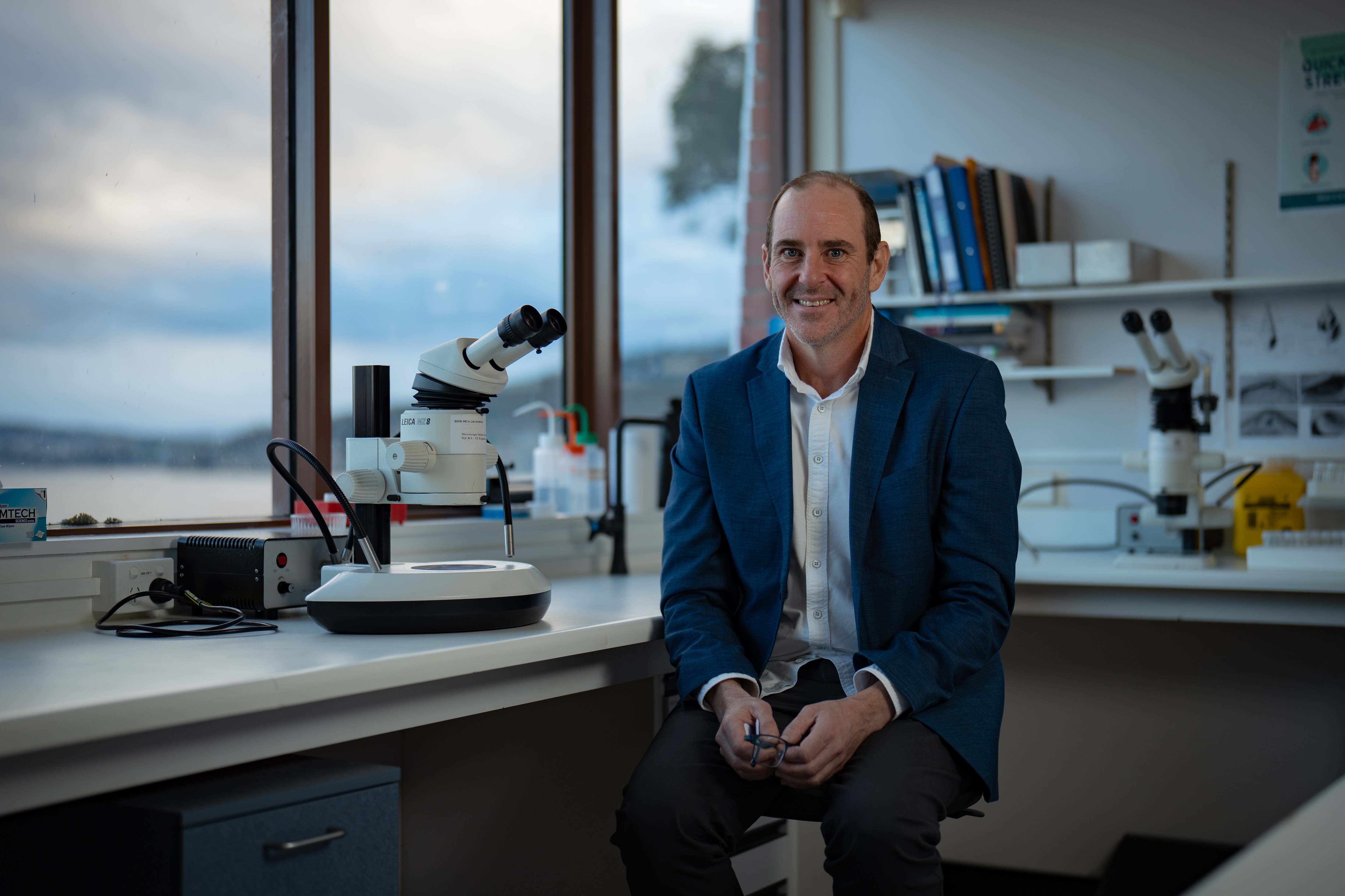 A man in a blue suit sits smiling in a science lab overlooking the ocean