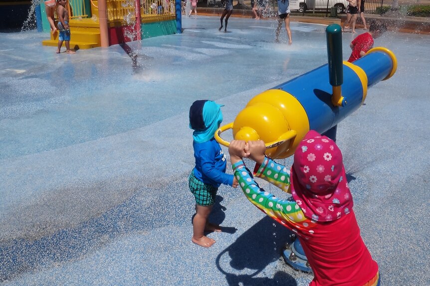 Two young children playing with a water cannon at a water park.  A fort with water fountains is in the background.