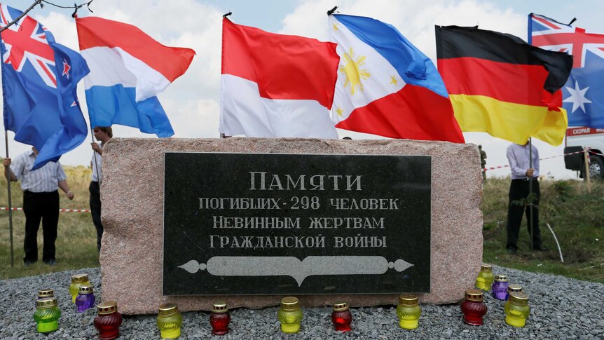 A large plaque in Ukranian marks the site of a downed Malaysian Airlines flight as people hold national flags behind it.