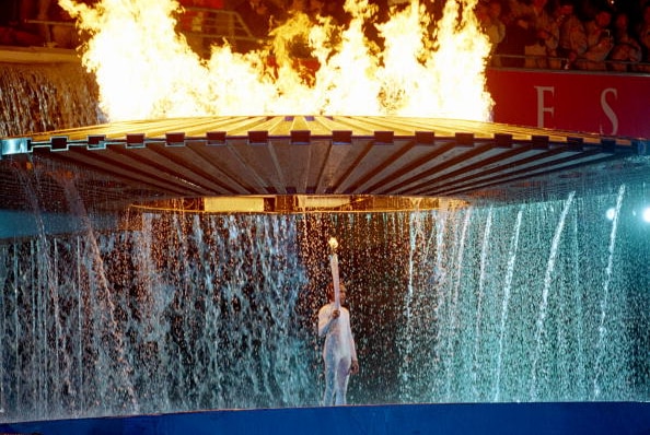 A woman stands with a flaming torch, as a large ring of fire rises behind her with water cascading off it