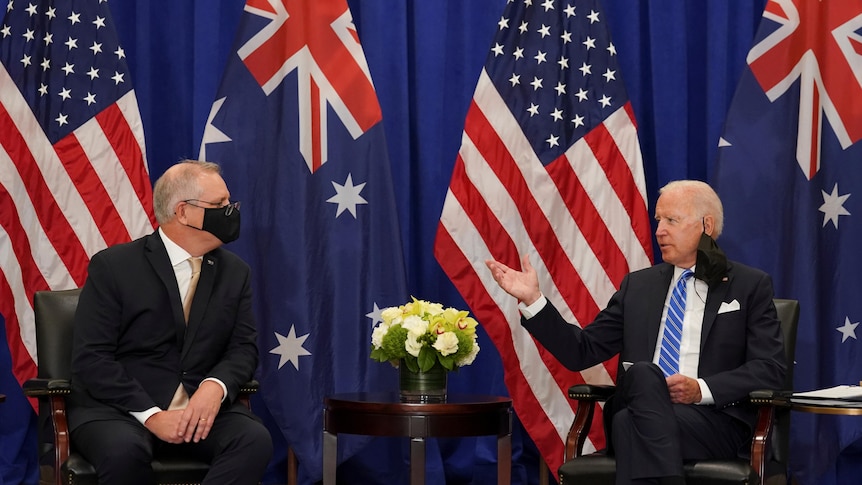 Two suited men sit and talk in front of two Australian and two US flags.