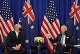 Two suited men sit and talk in front of two Australian and two US flags.