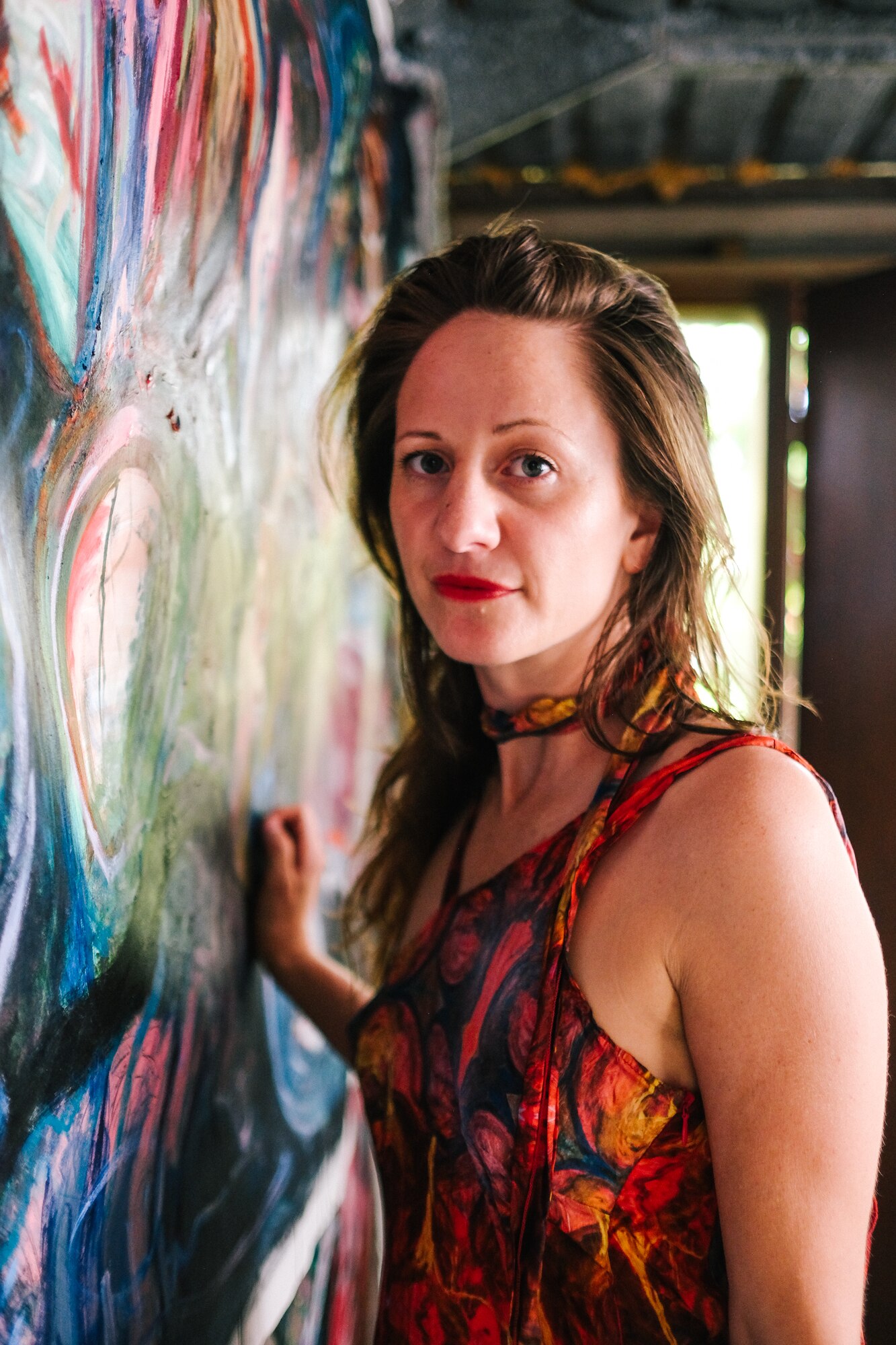Photo of artist Nara Walker standing close to a painting and looking at the camera. She has long brown hair and red lipstick