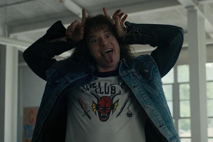 Un adolescente con una camiseta del Hellfire Club con un diablo y una chaqueta vaquera hace cuernos de diablo en la cabeza.