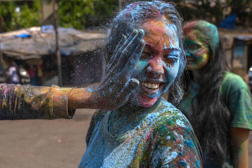 A smiling woman is covered in brightly coloured powders as a hand on the left side applies powder on her face.