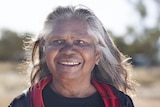 An older Indigenous woman wearing a black shirt smiles into the distance in a bush setting