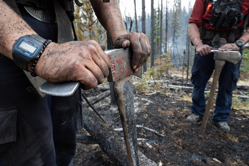 Close-ups of two axes being leaned on by dirty hands in a forest.