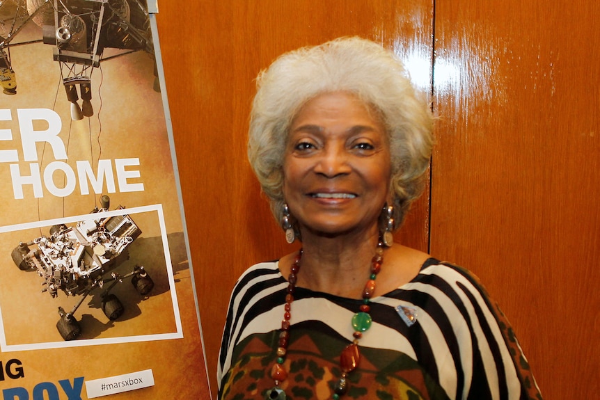 Actor Nichelle Nichols poses in front of an orange wall and a NASA recruitment sign. 