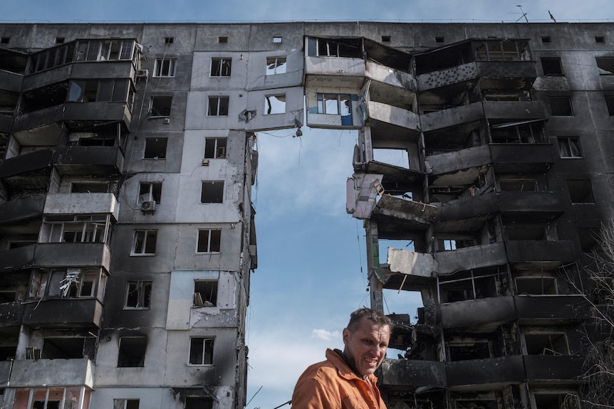 A man stands in from of a large damaged building, in which many walls are missing.