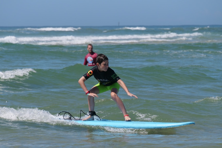 Boy aged about 10 surfs small wave standing up with knees bent as surf coach watches in background.