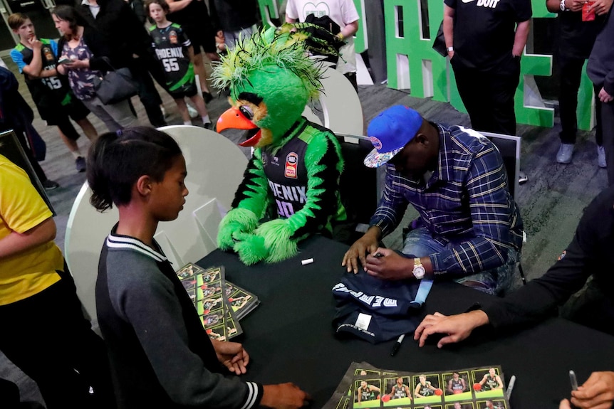 Zach Randolph uses his left hand to sign a t-shirt while sitting at a table, as a young fan looks on.