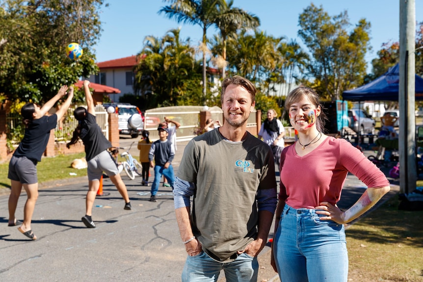 An image of a man and woman standing on a street with children playing volleball in the background