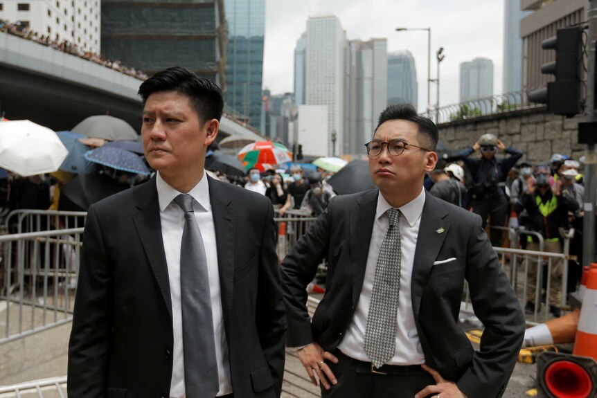 Two men in suits stand on the streets of Hong Kong with protestors holding umbrellas in the background.