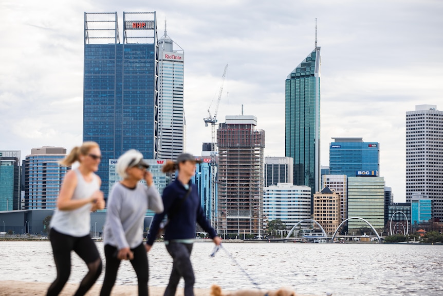Three women walk on a footpath along a river with a city skyline in the background.