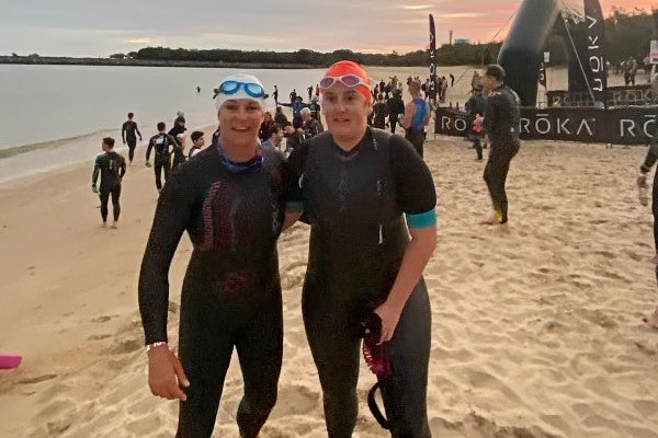 Two women in black wetsuits and swimming caps stand on a beach.