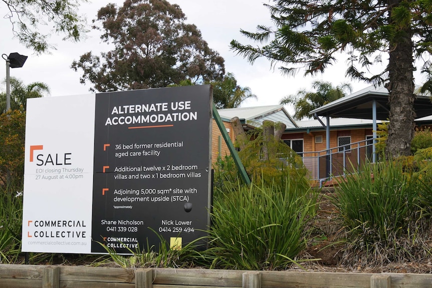 A large for sale sign detailing an aged care home facility in front of a building amongst shrubbery.