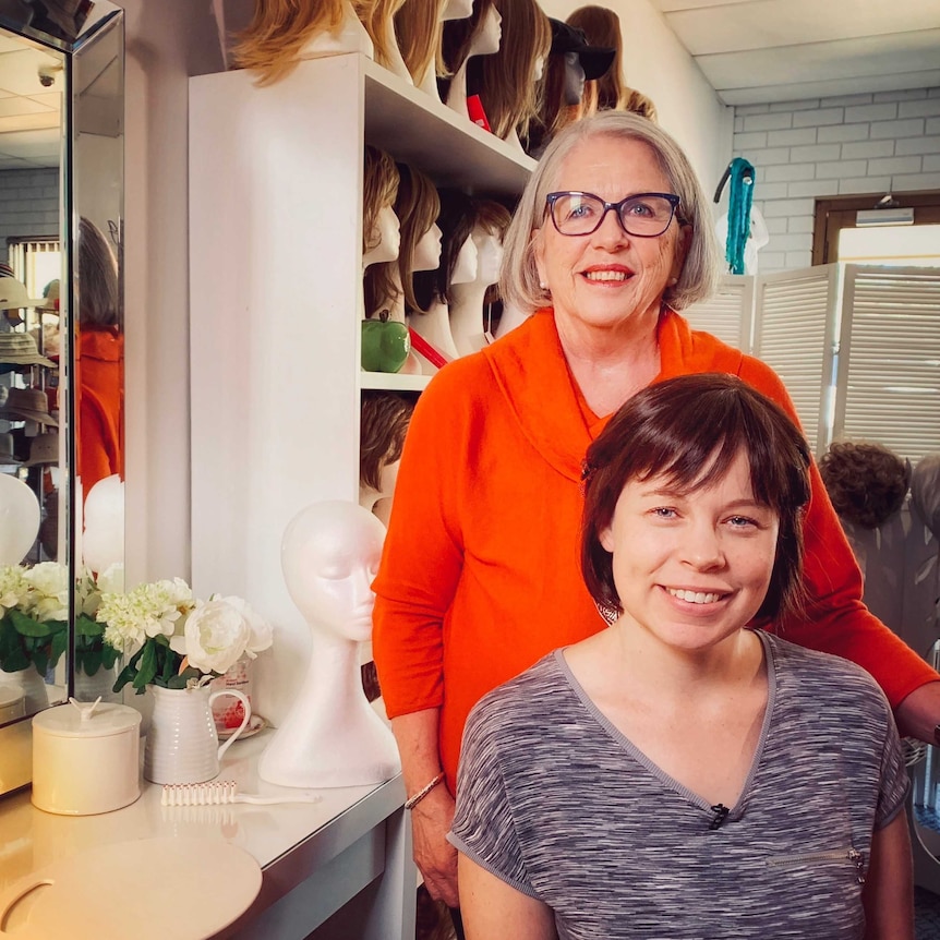 Two women look at the camera in a room full of wigs on stands.