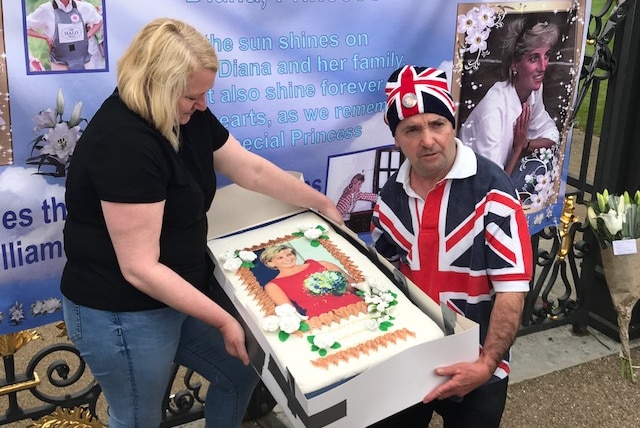 Maria Scott and John Loughrey hold a large cake with a photo of Princess Diana on it in front of the gates at Kensington Palace.