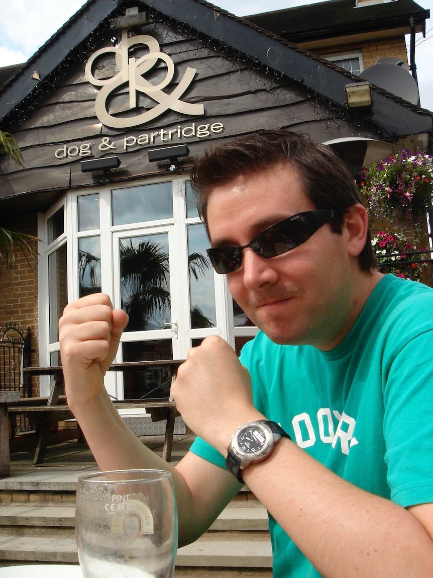 Michael Tetlow comically raises his fists while sitting at a table in front of the Dog and Partridge British pub.