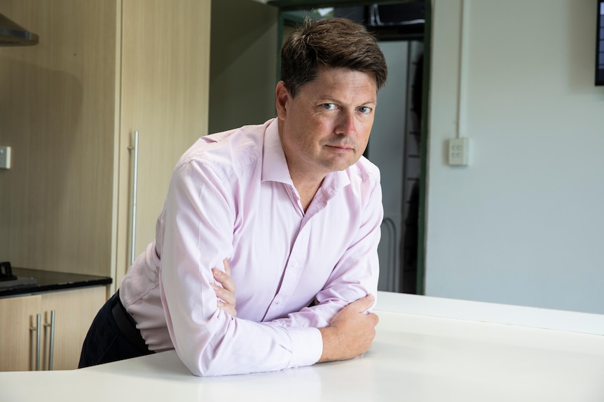 A man in a blue shirt and short brown hair leans against a table in a room that resembles a kitchen