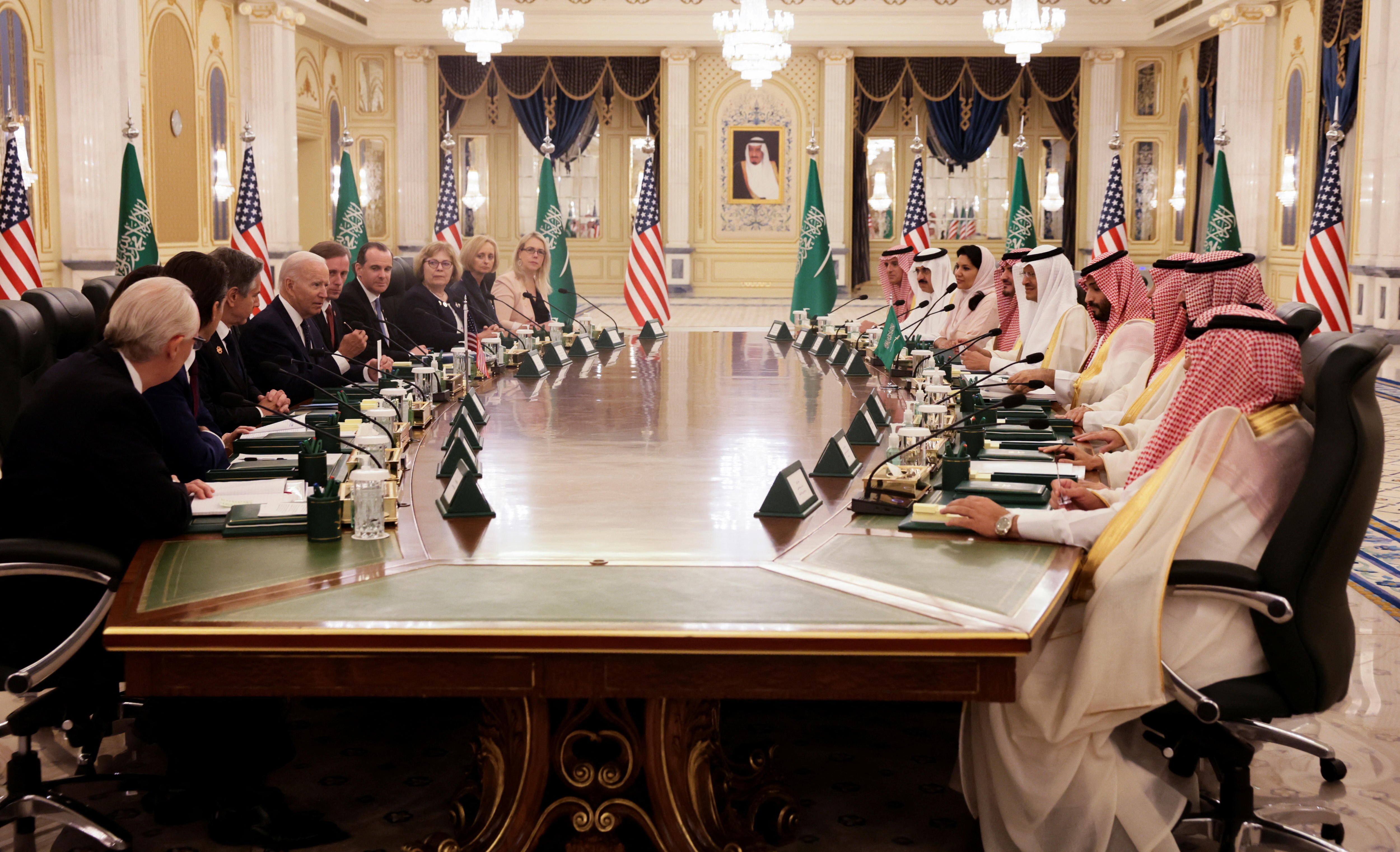 Two groups sit a cross from each other along a long table with many US and Saudi flags in the background.