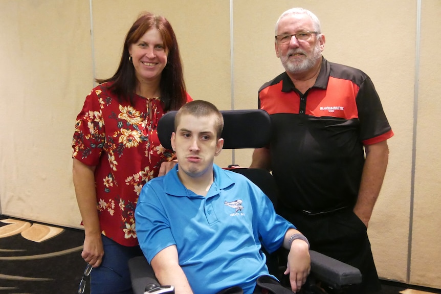A woman in a red shirt and man in a black and red shirt stand behind a boy in a wheelchair wearing a blue shirt.