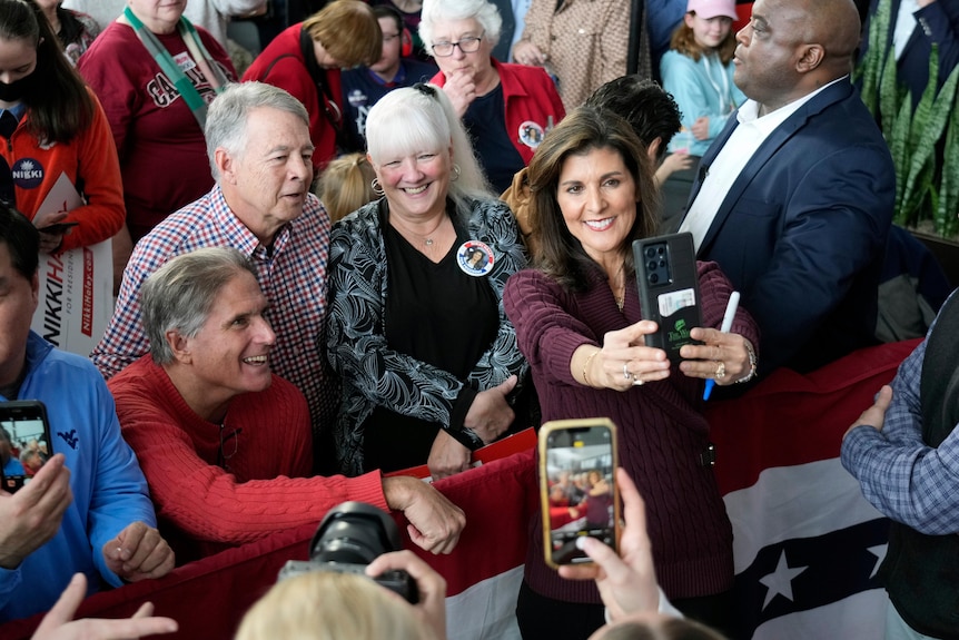 A middle-aged woman takes a selfie in front of a group of older voters in a crowd.