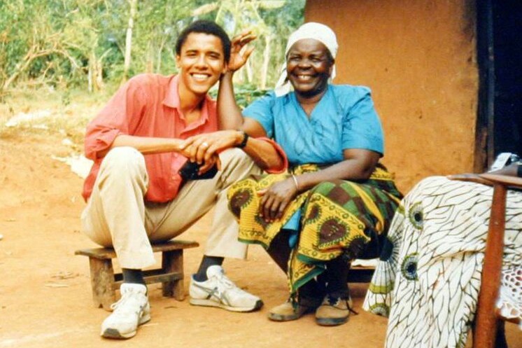  A young mixed-race man in a red shirt smiles and sits beside an older African woman in blue.