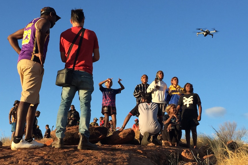 A group of children watch on adults fly a drone