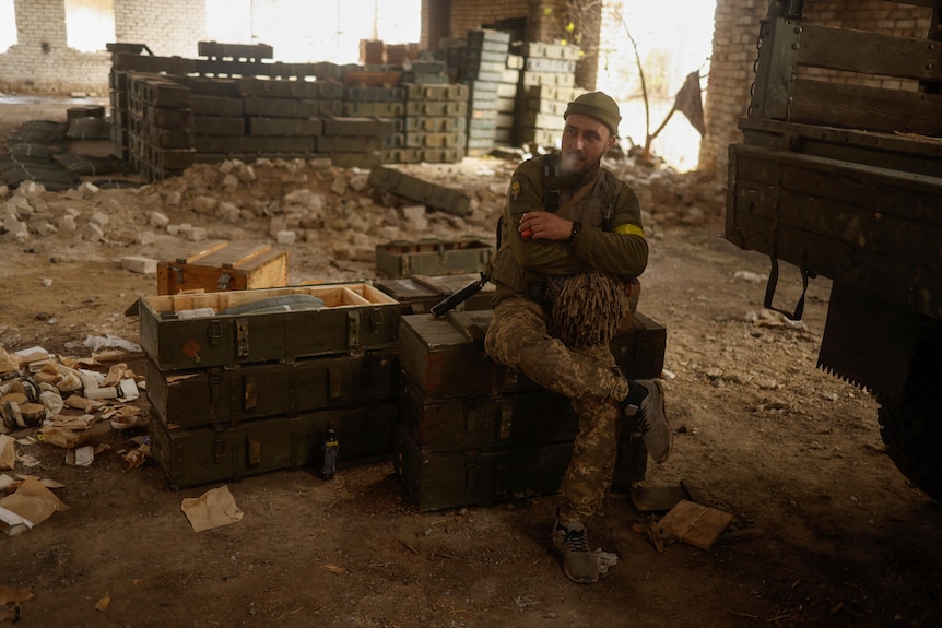 A soldier smoking sits on green wooden boxes in a dirt floor room. More boxes are piled high behind him. 