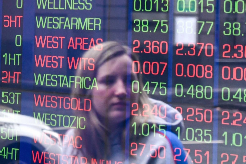 A woman is seen in the reflection of a window looking at shares' performance on a board of the ASX