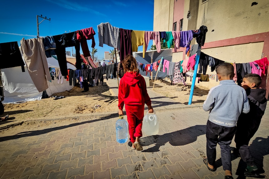 A little girl walks with two big jugs of water through a camp