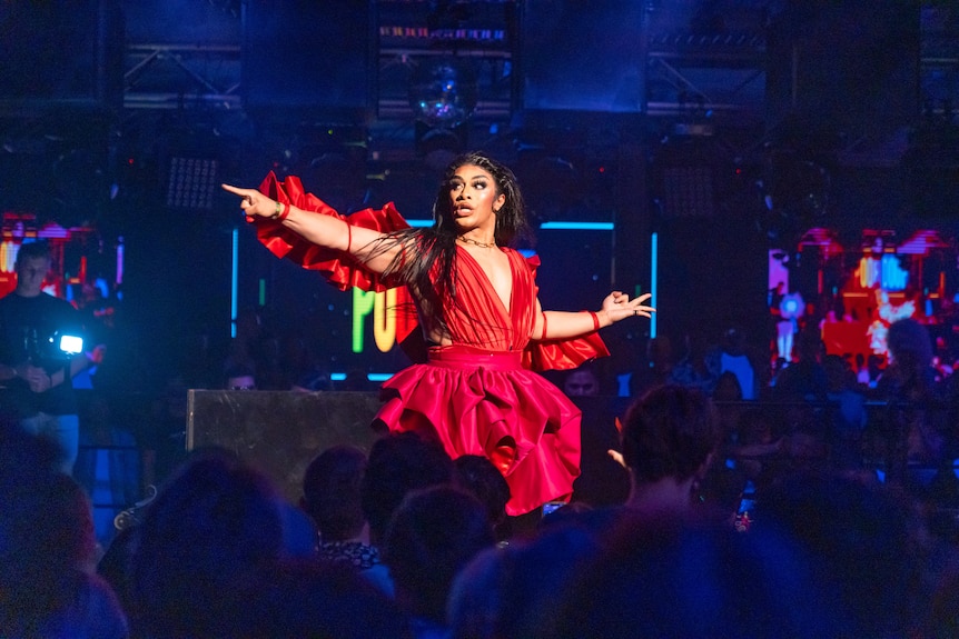 Kween Kong, in a red dress, points to the side of the stage.