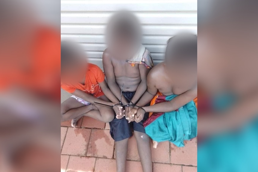 Three children sit with their hands cable tied. Their faces are blurred. 