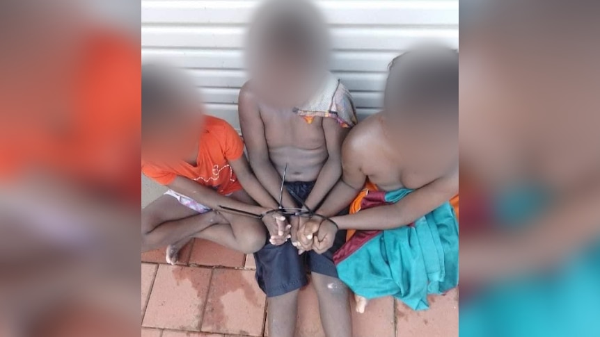 Three children sit with their hands cable tied. Their faces are blurred. 