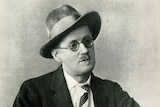 A black and white image of a man wearing a hat, glasses and a suit and tie.